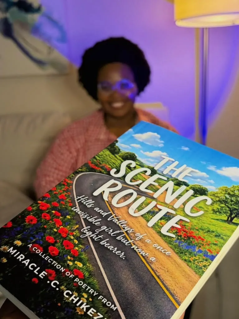 A copy of The Scenic Route held towards the camera, Miracle smiling behind it.