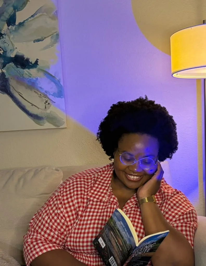 Miracle in a red gingham shirt, smiling as she reads The Scenic Route on a sofa.